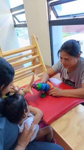 La composición de la imagen está centrada en la interacción entre una adulta y un niño pequeño, con otra persona que parece estar asistiendo o supervisando la actividad. Se aprecia un juego de anillos apilables sobre una superficie roja. El encuadre es de plano medio, permitiendo ver a los participantes y algunos elementos del entorno.