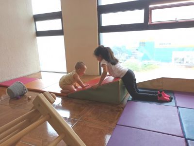 La imagen captura un momento de juego entre dos niños en un espacio interior, donde una niña observa a un bebé que está gateando sobre un pequeño objeto para trepar.