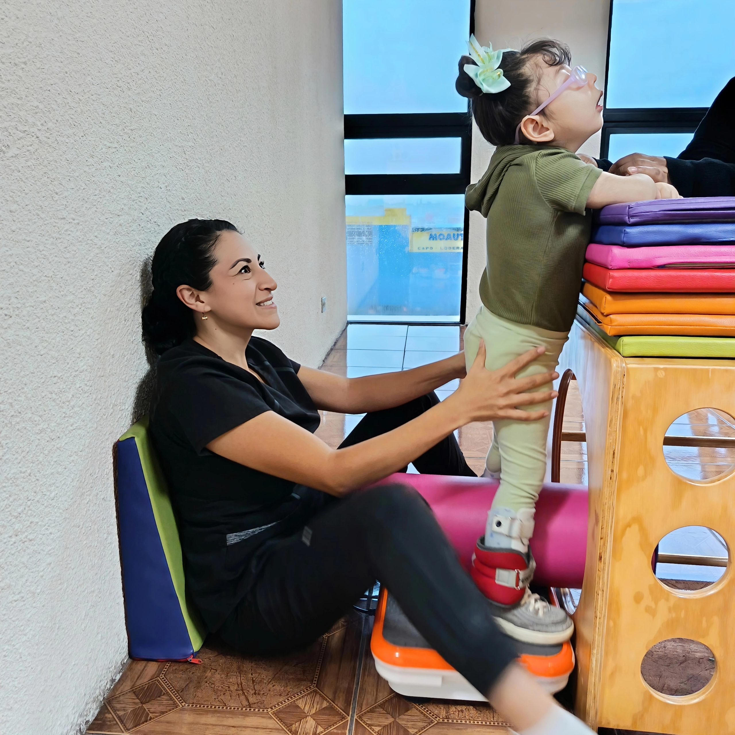 Una imagen conmovedora muestra a una terapeuta ayudando a una niña a superar una barrera de juegos, en un momento de apoyo y progreso. La escena está compuesta de manera que captura la interacción entre la terapeuta y la niña. La terapeuta está sentada en el suelo, apoyada contra una pared, y extiende sus manos para apoyar a la niña, quien está subiendo sobre una estructura de madera con coloridos bloques encima. La niña está mirando hacia arriba, posiblemente enfocándose en su objetivo, y se inclina hacia adelante. La composición muestra un momento de colaboración y aliento.